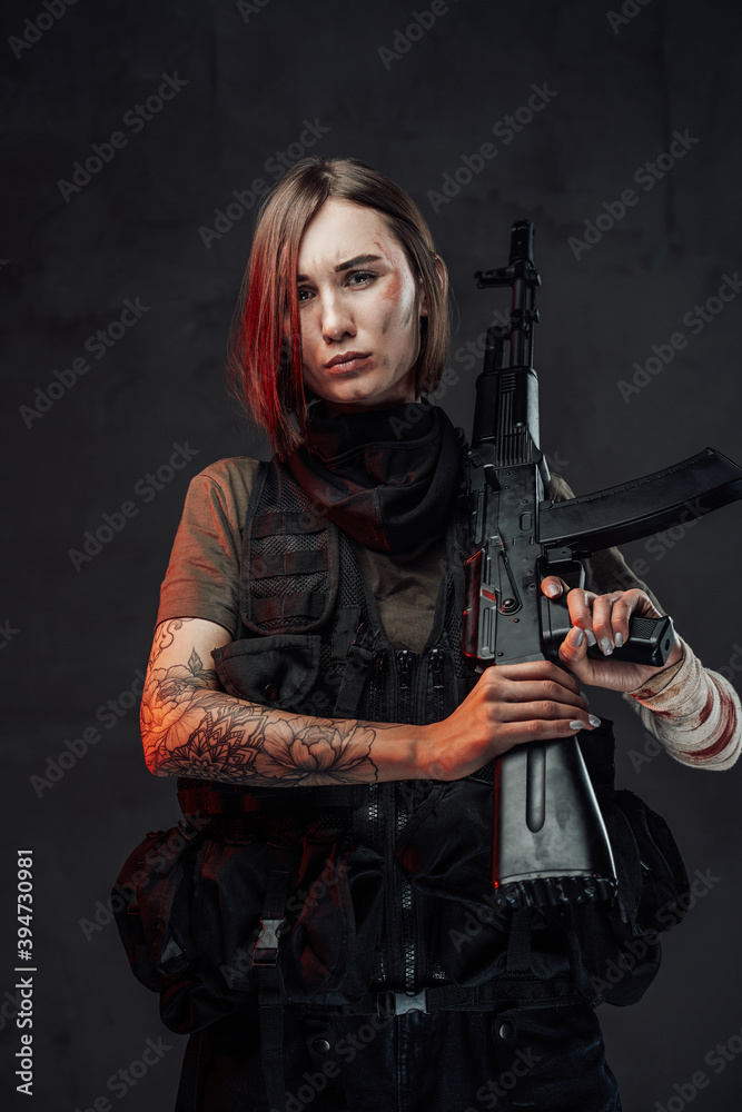 Dreseed in dark armour military woman with short haircut holds rifle with tattooed and bandaged hands in dark background.