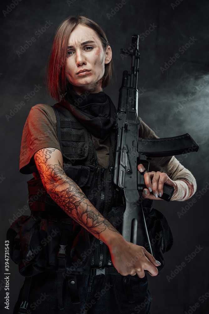 Dramatic portrait of tattooed female soldier with bandaged hand and short haircut poses holding rifle on her shoulder in dark background.