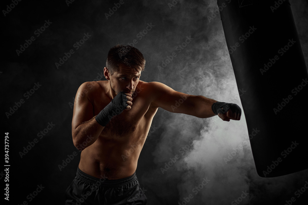 Aggressive shirtless boxer training defense and attacks in boxing bag on black background with smoke