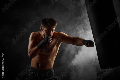 Aggressive shirtless boxer training defense and attacks in boxing bag on black background with smoke