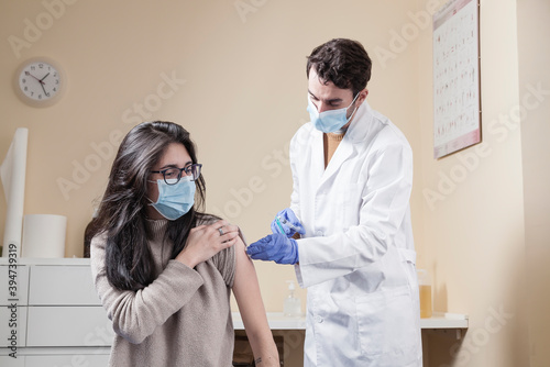 White woman with long hair wearing protective mask and glasses looking to the doctor speaking with him while being vaccinated on a clinic. Vaccine for the pandemics concept.