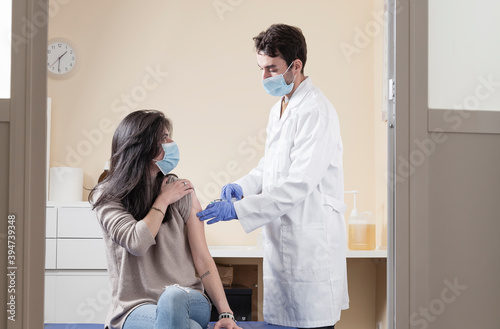 White woman with long hair wearing protective mask looking to the doctor speaking with him while being vaccinated on a clinic. Vaccine for the pandemics concept with door frame negative space.