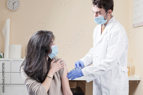 White woman with long hair wearing protective mask looking to the doctor smiling, trusting and speaking with him while being vaccinated on a clinic. Vaccine for the pandemics concept.