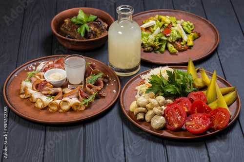 Food set with grilled pork ribs, pickled vegetables, bacon, salad, and moonshine on black wooden background
