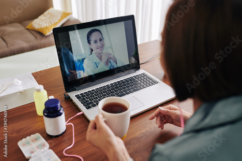 Young woman drinking cup of hot tea when sitting at table with laptop and video calling her doctor