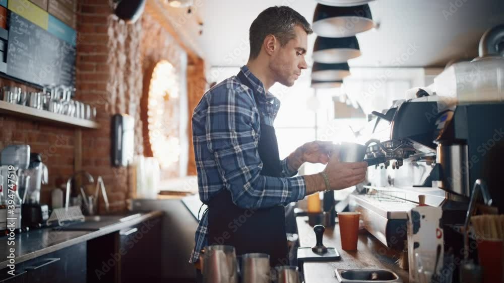 Handsome Male Barista in Checkered Shirt is Making a Take Away Cappuccino in Disposable Recycle Cup for a Customer in a Coffee Shop Bar. Female Cashier Works at a Cozy Cafe Counter in the Background. Handsome Male Barista in Checkered Shirt is Making a Take Away Cappuccino in Disposable Recycle Cup for a Customer in a Coffee Shop Bar. Female Cashier Works at a Cozy Cafe Counter in the Background.