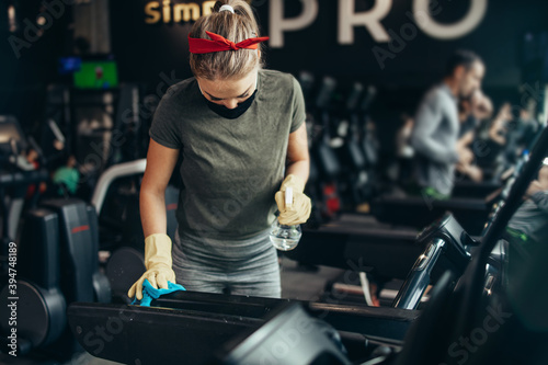 Young female worker disinfecting cleaning and weeping expensive fitness gym equipment with alcohol sprayer and cloth. Coronavirus global world pandemic and health protection safety measures. © Dusko