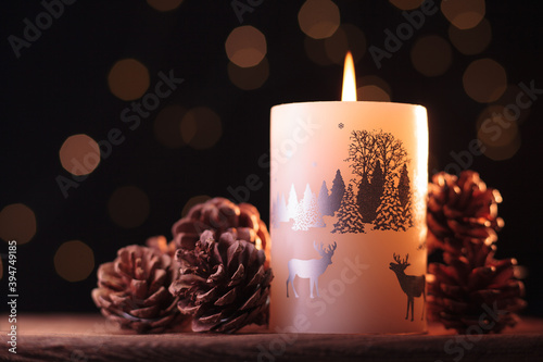 White Christmas candle and small brown pine cones on a wooden table with garlands background