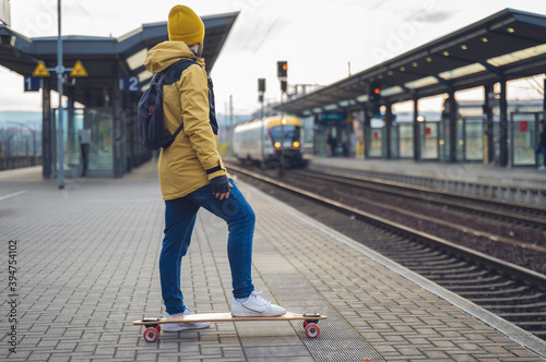 A boy, wearing winter clothes, skateboarding, is waiting for the train at the station. In the background the train arrives.