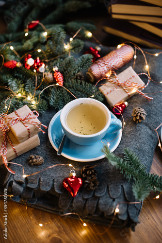 Delicious freshly brewed thyme tea in a blue cup standing on the warm cozy piled cover with red christmas ornamental hearts, little wrapped gifts, fireflies, pine cones on the wooden table background 