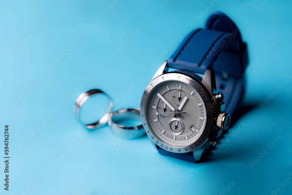 Reloj de pulsera plateado con correa azul sobre fondo azul cielo y anillos desenfocados con espacio para texto. Accesorios para hombre. Reloj de pulsera plateado con correa azul sobre fondo azul cielo y anillos desenfocados con espacio para texto. Accesorios para hombre.