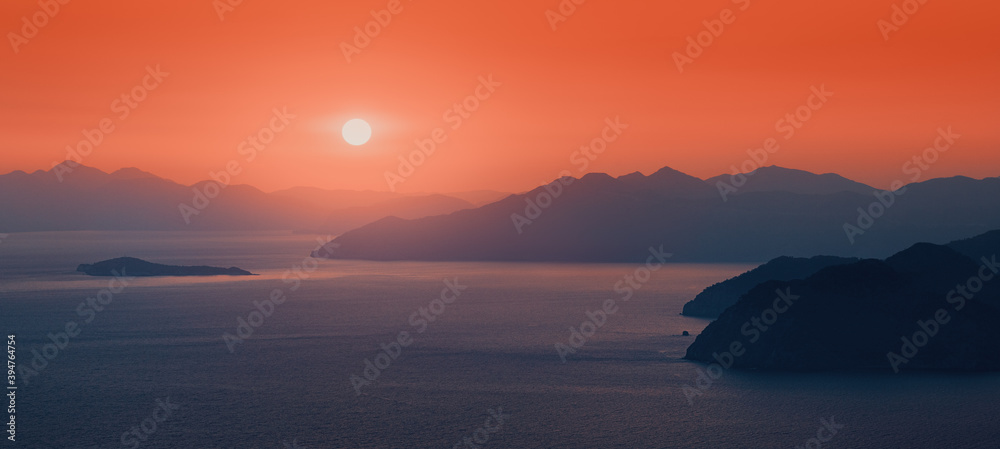 Fototapeta premium Moody sunset, the distant sun sets behind the silhouettes of blue mountains on the horizon, washed by the waters of the Mediterranean sea