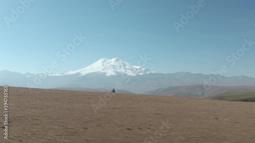 Person rides mtb bicycle along rural road in field against distant Elbrus mountain under clear sky on sunny day aerial bird eye view. photo