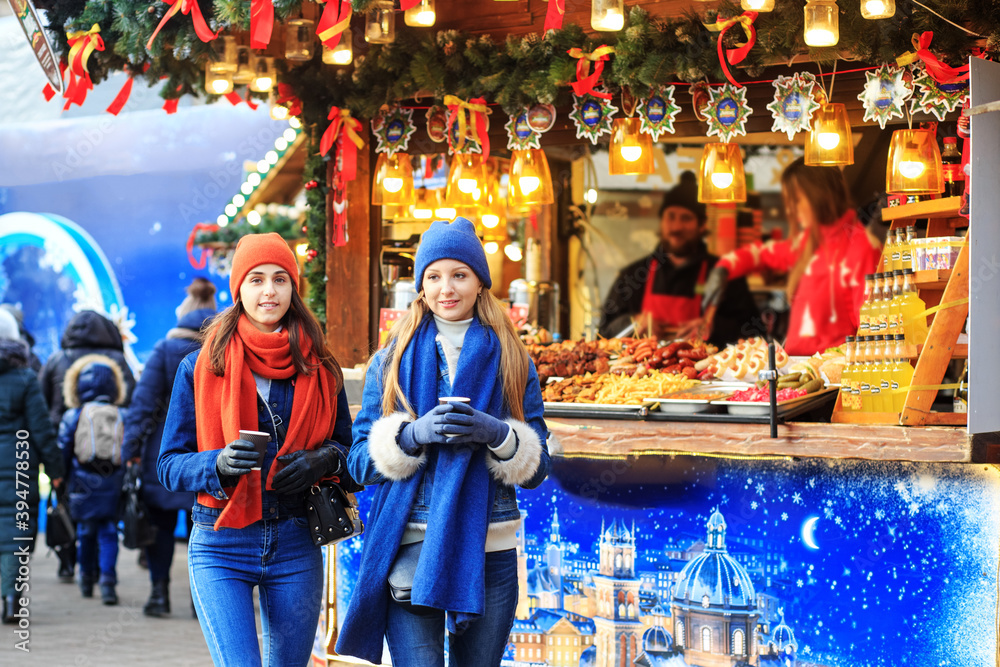 two young girls in red and blue hats and scarves walk around the Christmas market, drink coffee, talk and smile