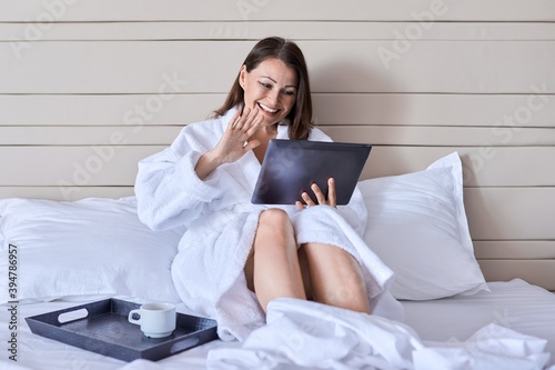 Mature female in white bathrobe with cup of coffee looking at webcam of digital tablet