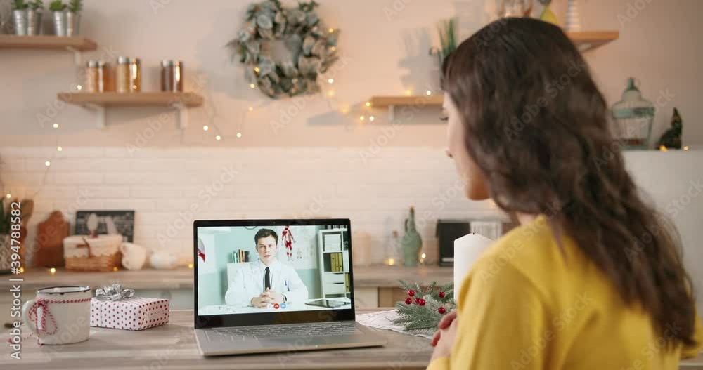 Over shoulder view. Close up of Caucasian woman indecorated home having online video call on laptop with male physician in quarantine in xmas festive period. Video consultation on computer on holidays Over shoulder view. Close up of Caucasian woman indecorated home having online video call on laptop with male physician in quarantine in xmas festive period. Video consultation on computer on holidays