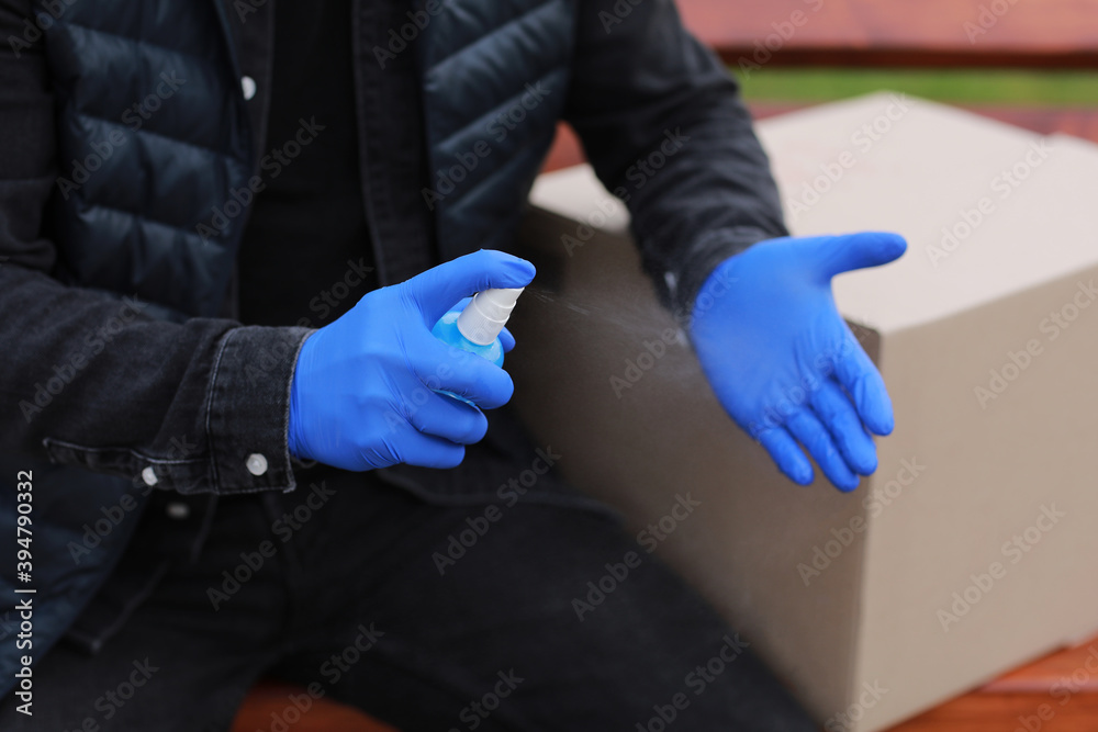 Delivery service courier during the Coronavirus, COVID-19, pandemic, cropped courier in medical protective mask, gloves spraying alcohol disinfectant spray on hands near cardboard boxes outdoors