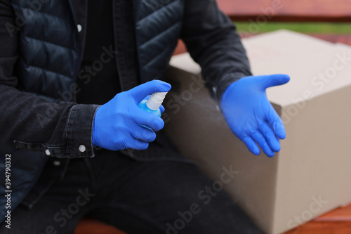 Delivery service courier during the Coronavirus, COVID-19, pandemic, cropped courier in medical protective mask, gloves spraying alcohol disinfectant spray on hands near cardboard boxes outdoors