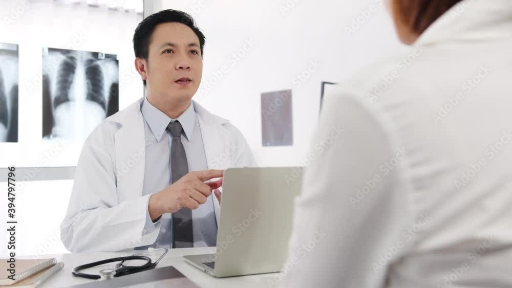 Serious Asia male doctor in white medical uniform using computer laptop is delivering great news talk discuss results or symptoms with female patient sit at desk in health clinic or hospital office. Serious Asia male doctor in white medical uniform using computer laptop is delivering great news talk discuss results or symptoms with female patient sit at desk in health clinic or hospital office.