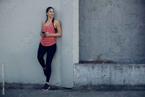 Attractive sportswoman leaning on the wall and using smart phone while pausing long run.