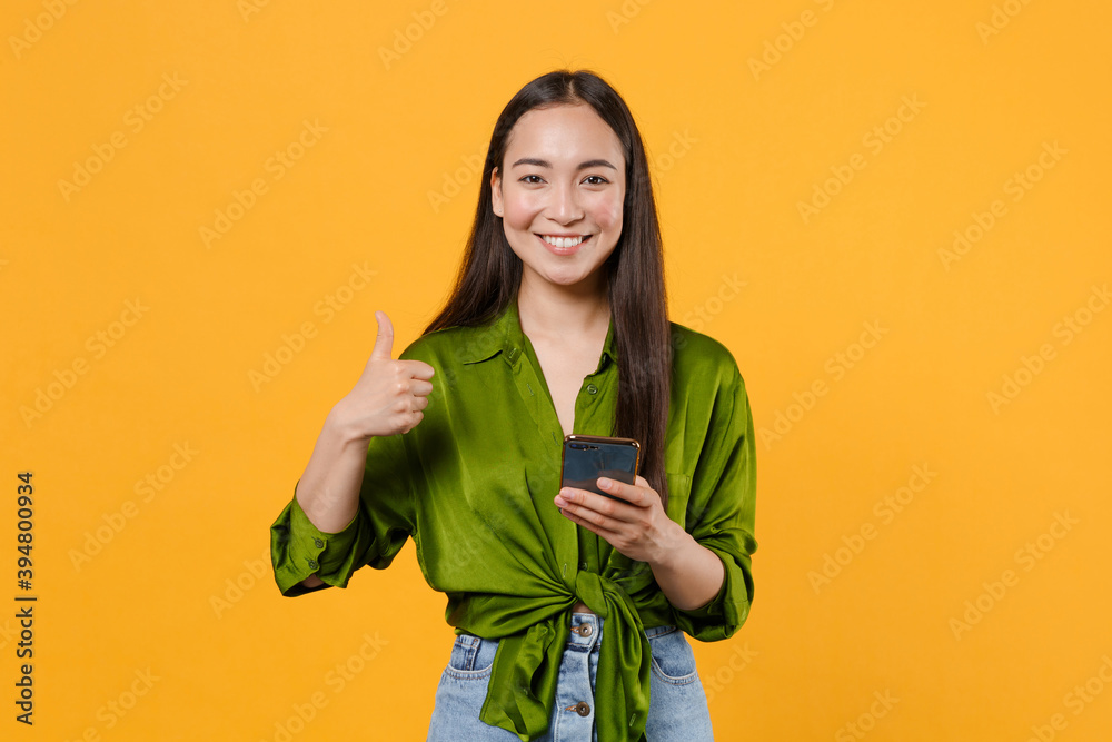 Smiling pretty young brunette asian woman wearing basic green shirt standing using mobile cell phone typing sms message showing thumb up isolated on bright yellow colour background, studio portrait. Smiling pretty young brunette asian woman wearing basic green shirt standing using mobile cell phone typing sms message showing thumb up isolated on bright yellow colour background, studio portrait.
