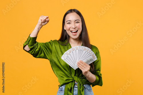 Fotografie Happy joyful young brunette asian woman wearing basic green shirt standing hold fan of cash money in dollar banknotes doing winner gesture isolated on bright yellow colour background, studio portrait