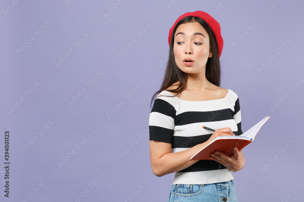 Shocked amazed young brunette asian student woman 20s wearing striped t-shirt red beret standing writing notes in notebook looking aside isolated on pastel violet colour background studio portrait. Shocked amazed young brunette asian student woman 20s wearing striped t-shirt red beret standing writing notes in notebook looking aside isolated on pastel violet colour background studio portrait.