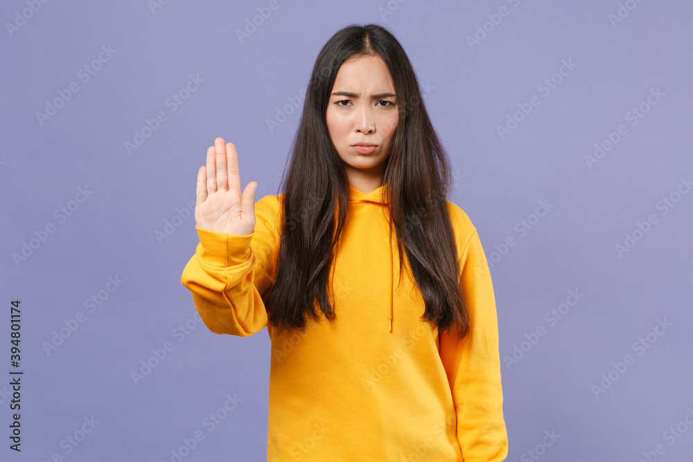 Dissatisfied displeased young brunette asian woman in casual basic yellow hoodie standing showing stop gesture with palm looking camera isolated on pastel violet colour background studio portrait. Dissatisfied displeased young brunette asian woman in casual basic yellow hoodie standing showing stop gesture with palm looking camera isolated on pastel violet colour background studio portrait.