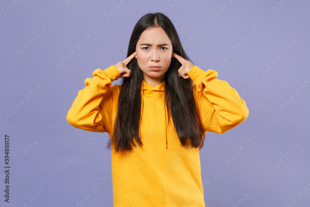 Dissatisfied displeased young brunette asian woman wearing casual basic yellow hoodie standing covering ears with fingers looking camera isolated on pastel violet colour background studio portrait. Dissatisfied displeased young brunette asian woman wearing casual basic yellow hoodie standing covering ears with fingers looking camera isolated on pastel violet colour background studio portrait.