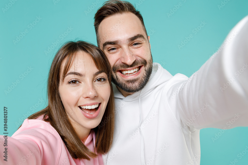 Close up of cheerful young couple two friends man woman 20s in white pink casual hoodie standing doing selfie shot on mobile phone isolated on blue turquoise colour wall background studio portrait. Close up of cheerful young couple two friends man woman 20s in white pink casual hoodie standing doing selfie shot on mobile phone isolated on blue turquoise colour wall background studio portrait.
