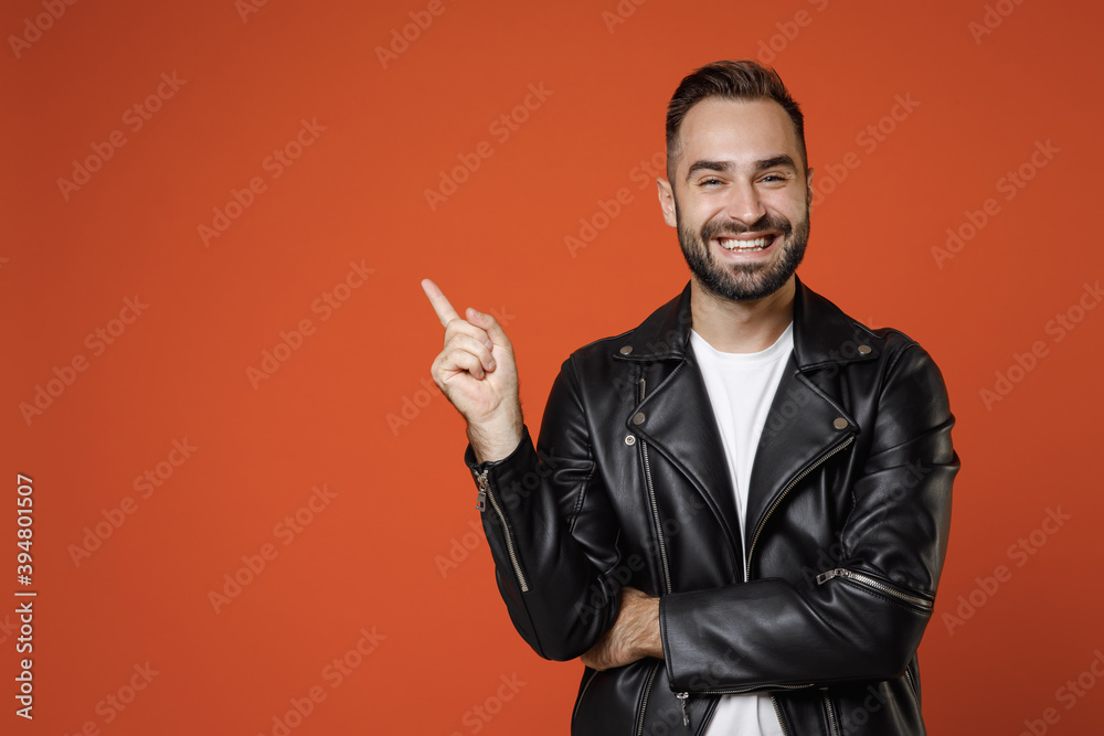 Smiling funny young bearded man wearing basic white t-shirt black leather jacket standing pointing index finger up on mock up copy space isolated on bright orange colour background studio portrait. Smiling funny young bearded man wearing basic white t-shirt black leather jacket standing pointing index finger up on mock up copy space isolated on bright orange colour background studio portrait.