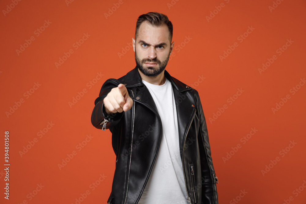 Strict irritated dissatisfied young bearded man wearing basic white t-shirt black leather jacket standing pointing index finger on camera isolated on bright orange colour background studio portrait. Strict irritated dissatisfied young bearded man wearing basic white t-shirt black leather jacket standing pointing index finger on camera isolated on bright orange colour background studio portrait.