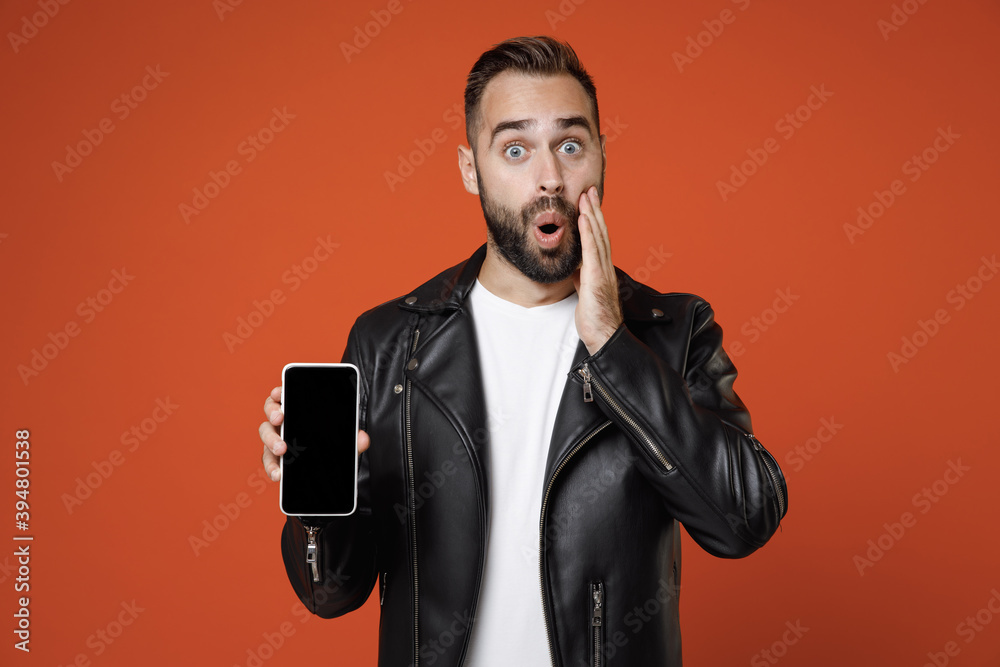 Shocked young bearded man 20s in white t-shirt black leather jacket put hand on cheek hold mobile cell phone with blank empty screen mock up copy space isolated on orange background studio portrait. Shocked young bearded man 20s in white t-shirt black leather jacket put hand on cheek hold mobile cell phone with blank empty screen mock up copy space isolated on orange background studio portrait.