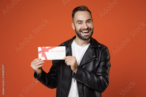 Cheerful funny young bearded man wearing basic white t-shirt, black leather jacket standing hold in hands gift certificate looking camera isolated on bright orange colour background studio portrait.