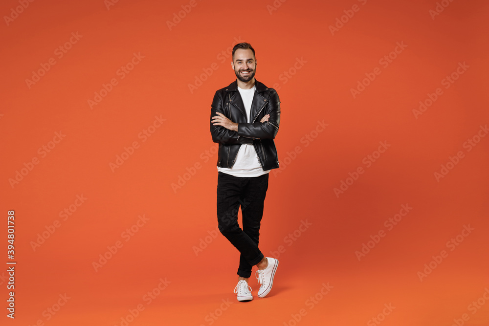 Full length of smiling attractive young bearded man 20s wearing basic white t-shirt black leather jacket standing holding hands crossed looking camera isolated on orange background studio portrait. Full length of smiling attractive young bearded man 20s wearing basic white t-shirt black leather jacket standing holding hands crossed looking camera isolated on orange background studio portrait.