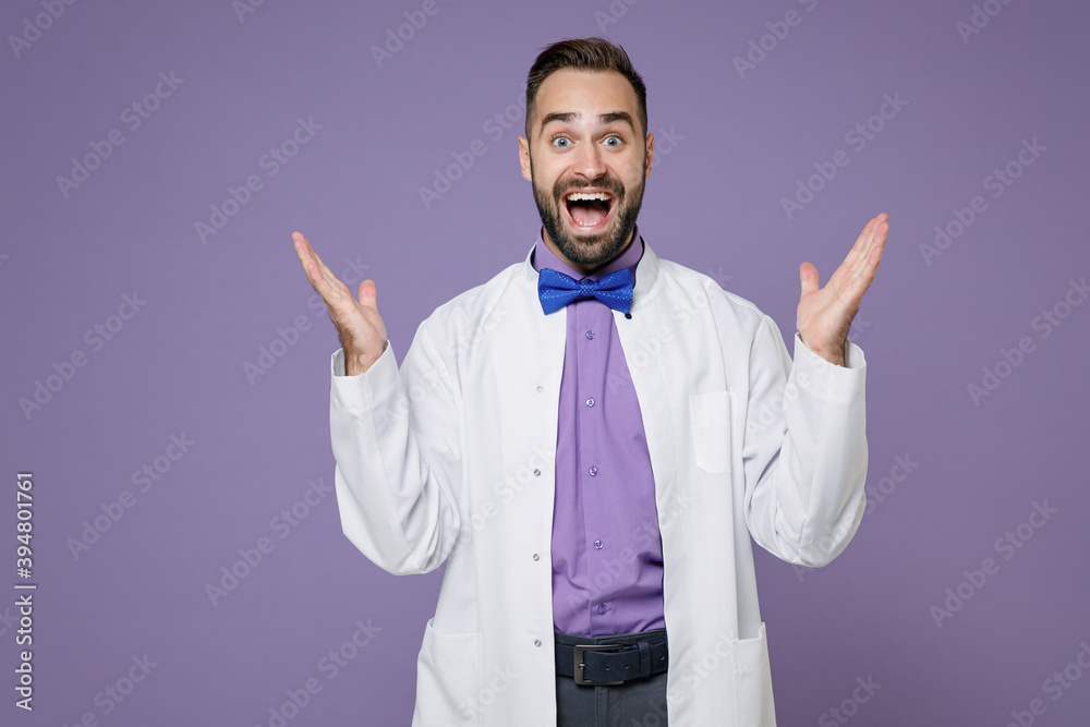 Surprised young bearded doctor man wearing white medical gown keeping mouth open spreading hands isolated on violet colour background studio portrait. Healthcare personnel health medicine concept. Surprised young bearded doctor man wearing white medical gown keeping mouth open spreading hands isolated on violet colour background studio portrait. Healthcare personnel health medicine concept.