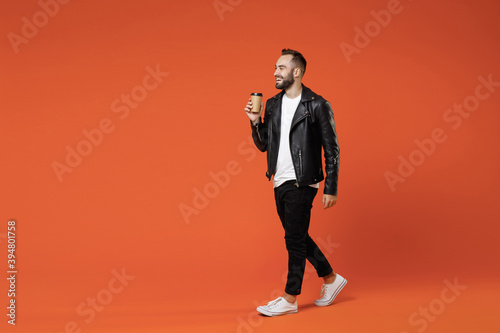 Full length side view smiling young bearded man in basic white t-shirt black leather jacket standing hold paper cup of coffee or tea looking aside isolated on orange colour background studio portrait. © ViDi Studio