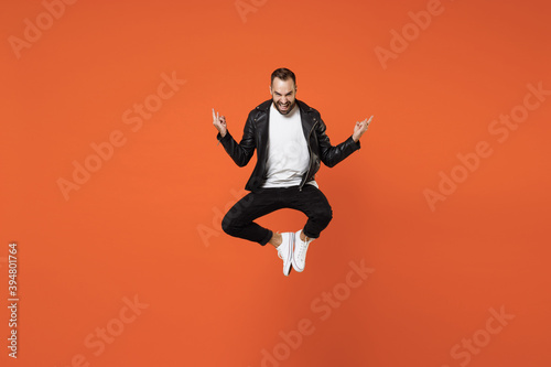 Full length of cheerful young bearded man 20s in basic white t-shirt black leather jacket jumping depicting heavy metal rock sign looking camera isolated on orange colour background studio portrait.