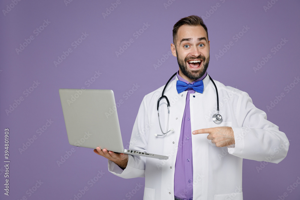 Excited young bearded doctor man in white medical gown stethoscope point index finger on laptop pc computer isolated on violet background studio portrait. Healthcare personnel health medicine concept. Excited young bearded doctor man in white medical gown stethoscope point index finger on laptop pc computer isolated on violet background studio portrait. Healthcare personnel health medicine concept.
