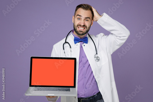 Puzzled young doctor man in medical gown stethoscope working on laptop computer with blank empty screen put hand on head isolated on violet background. Healthcare personnel health medicine concept.