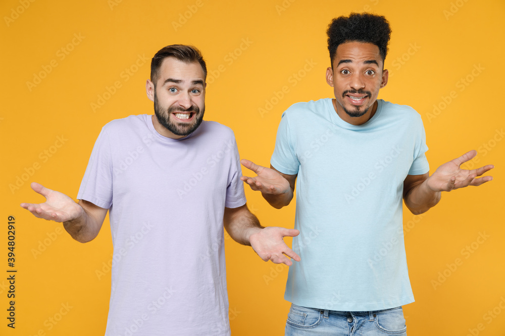 Confused puzzled young friends european african american men 20s wearing casual violet blue t-shirts spreading hands looking camera isolated on bright yellow colour wall background studio portrait. Confused puzzled young friends european african american men 20s wearing casual violet blue t-shirts spreading hands looking camera isolated on bright yellow colour wall background studio portrait.
