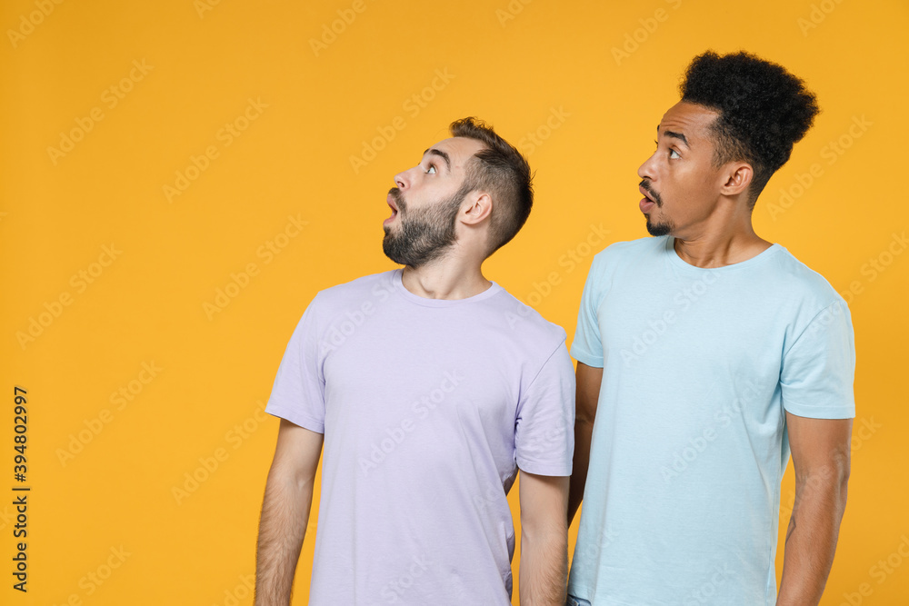 Shocked amazed young two friends european african american men 20s in violet blue casual t-shirts looking aside up keeping mouth open isolated on bright yellow colour wall background studio portrait. Shocked amazed young two friends european african american men 20s in violet blue casual t-shirts looking aside up keeping mouth open isolated on bright yellow colour wall background studio portrait.