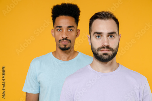 Handsome serious young two friends european african american men 20s wearing violet blue casual t-shirts standing and looking camera isolated on bright yellow colour wall background studio portrait.
