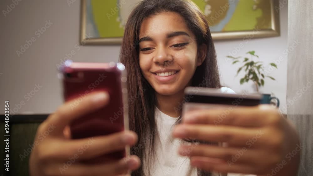 Smiling young african american woman with credit card and smartphone sitting on couch at home. Happy shopper using instant easy mobile payments making purchase in online store. Smiling young african american woman with credit card and smartphone sitting on couch at home. Happy shopper using instant easy mobile payments making purchase in online store.