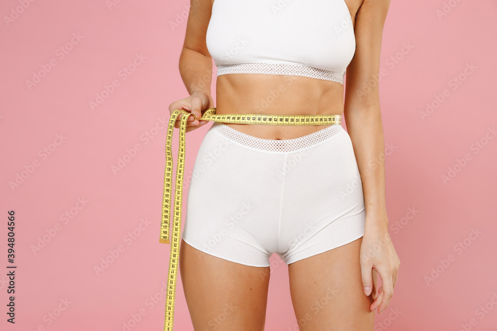 Cropped image of strong fitness sports young woman 20s in white underwear with strong body posing measuring waist with measure tape isolated on pastel pink colour wall background, studio portrait. - obrazy, fototapety, plakaty 