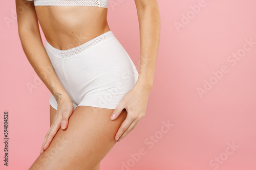 Cropped image side view of feminine young woman 20s wearing white brassiere underwear with sexy beautiful body standing posing hold hands on legs isolated on pastel pink background, studio portrait.