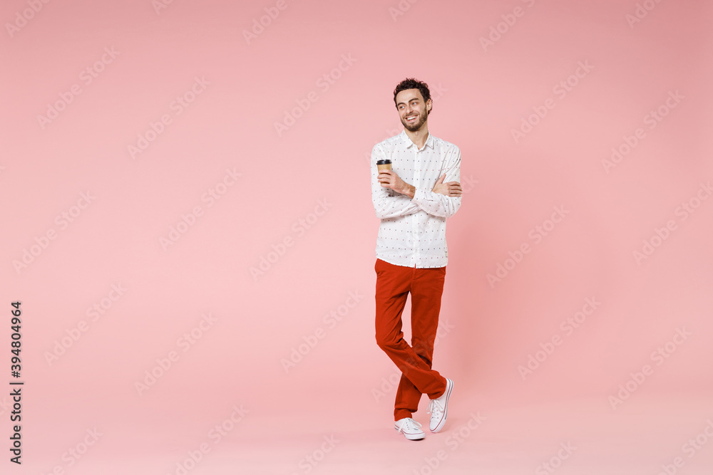 Full length of cheerful young bearded man 20s wearing basic casual white shirt standing hold paper cup of coffee or tea looking aside isolated on pastel pink color wall background studio portrait. Full length of cheerful young bearded man 20s wearing basic casual white shirt standing hold paper cup of coffee or tea looking aside isolated on pastel pink color wall background studio portrait.