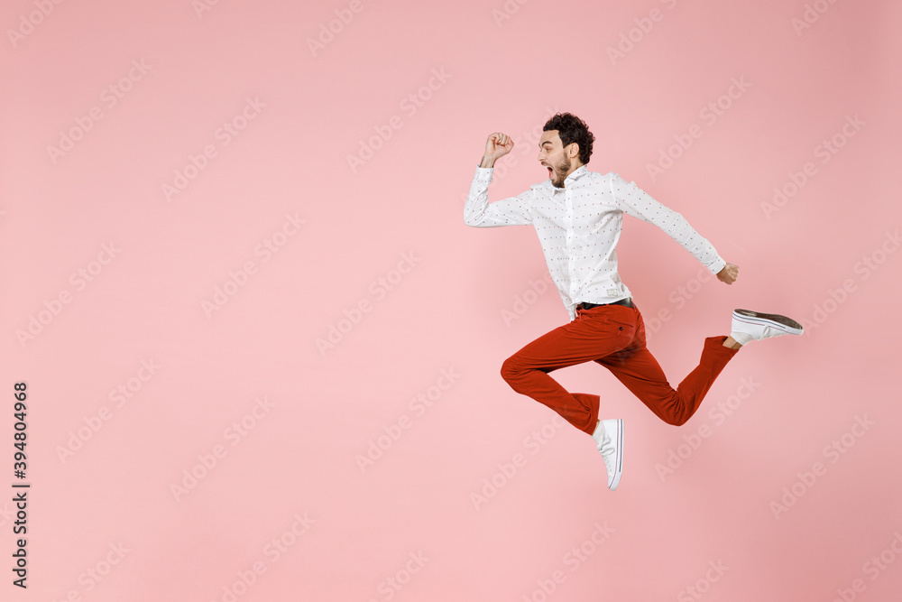 Full length side view of shocked screaming young bearded man 20s wearing basic casual white shirt jumping like running looking aside isolated on pastel pink color wall background studio portrait.