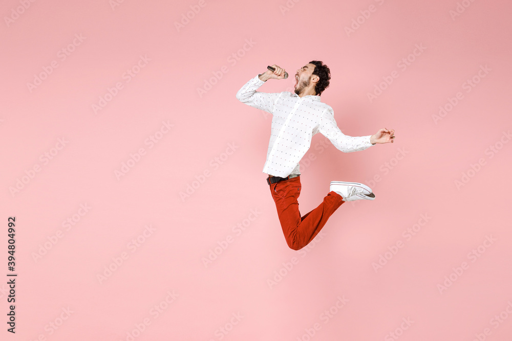 Full length side view of cheerful young bearded man 20s wearing basic casual white shirt jumping sing song in microphone spreading hands isolated on pastel pink color wall background studio portrait. Full length side view of cheerful young bearded man 20s wearing basic casual white shirt jumping sing song in microphone spreading hands isolated on pastel pink color wall background studio portrait.