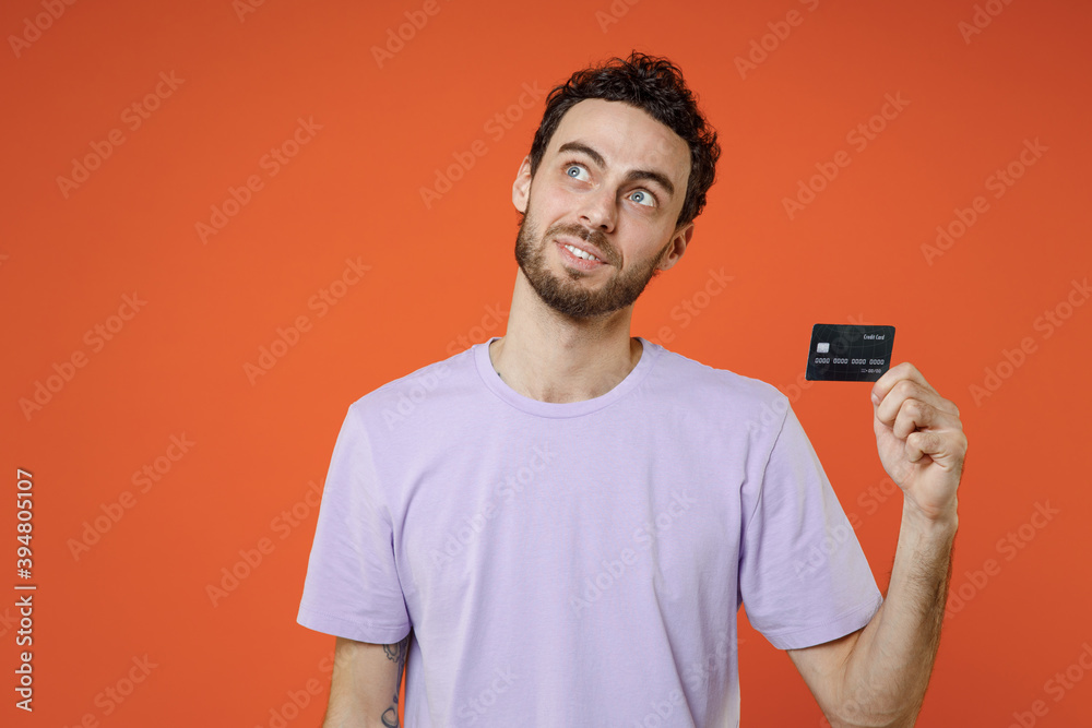 Smiling pensive young bearded man 20s wearing casual basic violet t-shirt standing holding in hand credit bank card looking aside up isolated on bright orange color wall background studio portrait. Smiling pensive young bearded man 20s wearing casual basic violet t-shirt standing holding in hand credit bank card looking aside up isolated on bright orange color wall background studio portrait.